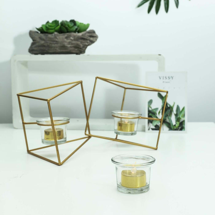 Gold geometric candle holders with a lit candle on a white table.
