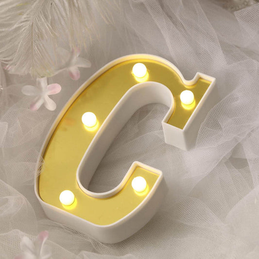 Gold letter "C" marquee light with white base on soft fabric background.