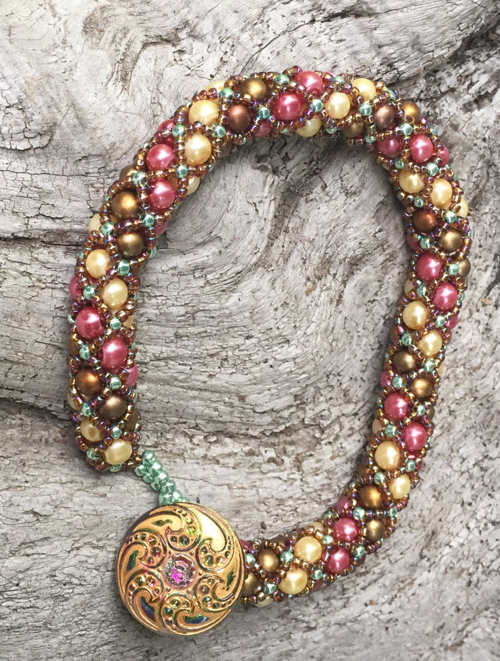 Netted Treasure style beaded bracelet with Swarovski Glass Pearls in Rose, Cream, and Matte Bronze, with Sage and Rootbeer seed beads holding the netting together. A subtly iridescent Czech Glass button closure with beaded loop finishes this work of art. This bracelet measures 8 1/4".