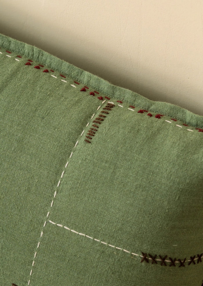 Close-up of a sage green fabric cushion or pillow with decorative hand-stitched seams in white, red, and dark brown.