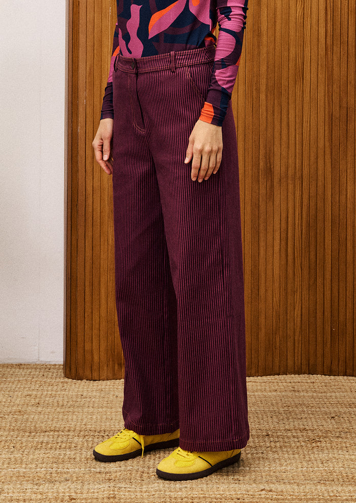 Person wearing a colorful top and purple corduroy pants with yellow shoes, standing against a wooden panel background.