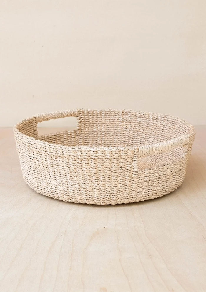 A round woven natural fiber basket with integrated side handles, shown on a light wood surface against a warm beige background.