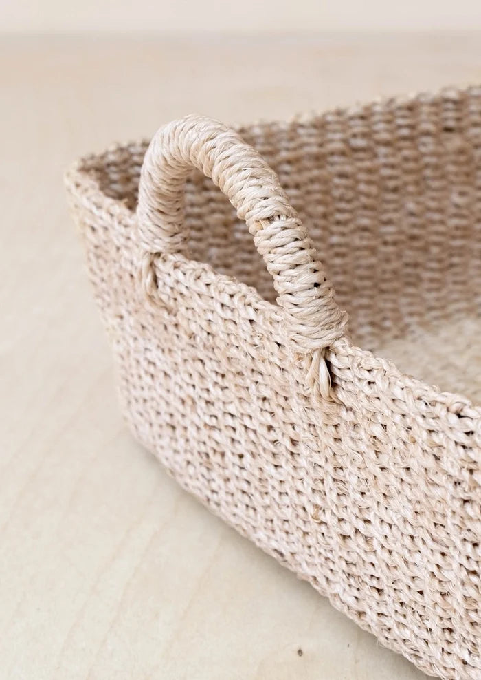 A close-up of a woven natural fiber storage basket with a single loop handle, shown on a light beige wooden surface.